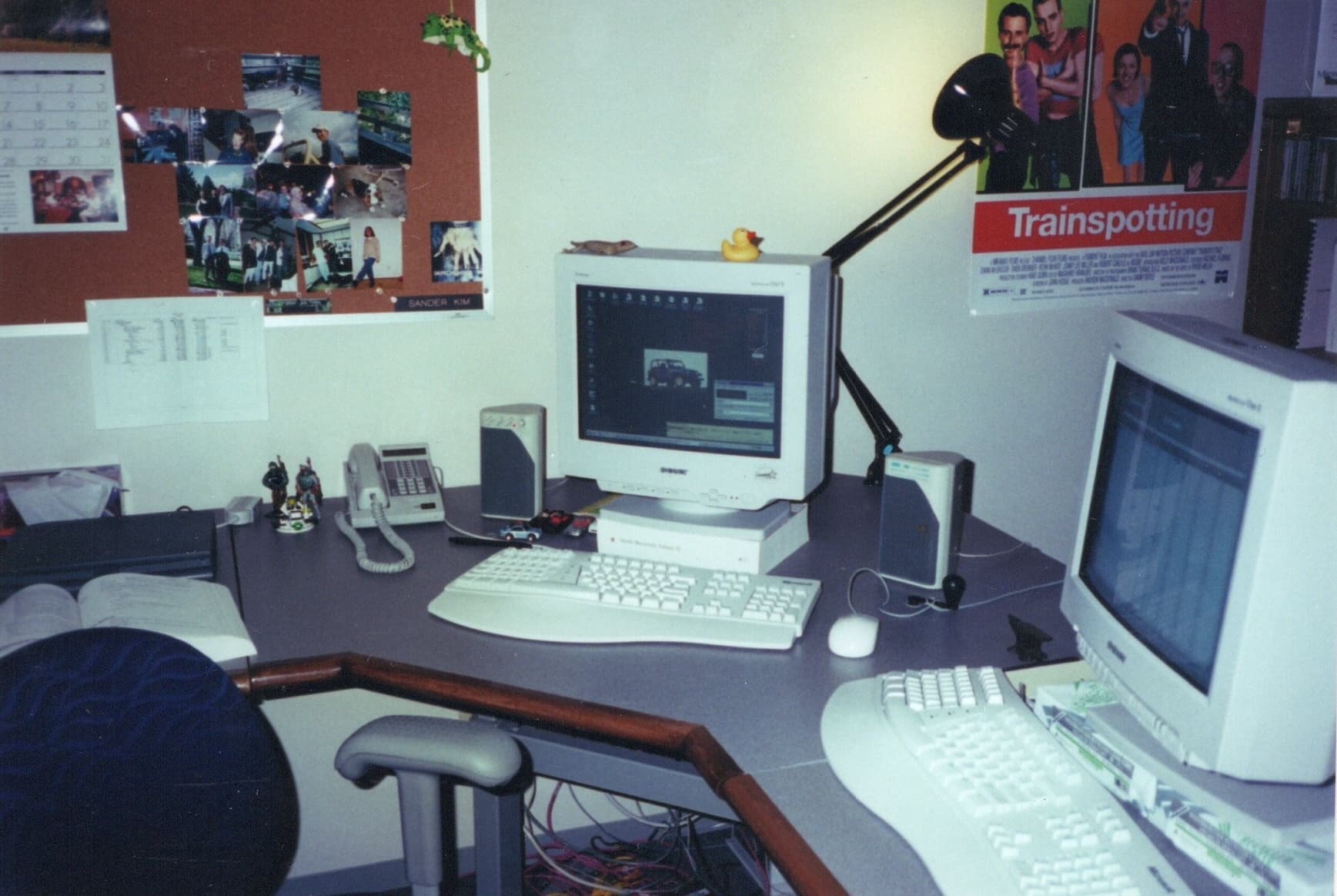 A Microsoft office desk with two CRT monitors, a phone, and a Trainspotting poster